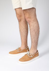 Beige Wildleder-Bootsschuhe mit einer weißen Gummisohle, die graue Schnürsenkel durch metallene Ösen gefädelt und ein lässiges Design aufweisen.