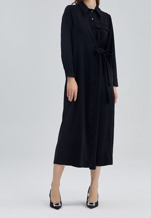 Femme portant une longue robe noire boutonnée avec une ceinture nouée à la taille et des talons noirs pointus ornés de boucles décoratives.