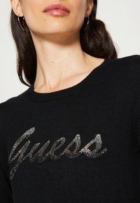 Fekete kötött pulóver, amelyet fényes "Guess" logó díszít strasszokkal, kerek nyakkivágással és puha tapintással.
