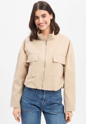 Lächelnde Frau mit schulterlangem dunklem Haar trägt eine beige Reißverschlussjacke mit großen Fronttaschen und blaue hochgeschnittene Jeans vor weißem Hintergrund.