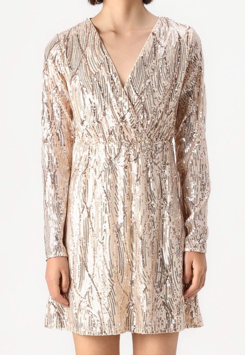 Femme portant une robe courte à manches longues et col en V, recouverte de sequins dorés et argentés, avec une taille cintrée et un motif abstrait en spirale.