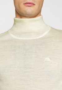 Cremefärgad polokrage tröja i fint stickat material, med en subtil broderad logotyp på bröstet. Slät yta med en figurnära siluett.