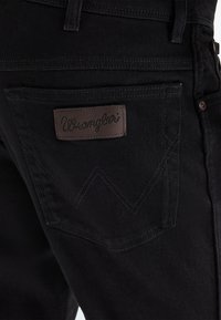 Záber na zadnú kapsu čiernych denimových džínsov s hnedou koženou nášivkou s nápisom „Wrangler“ a viditeľnými detailmi šitia kapsy.