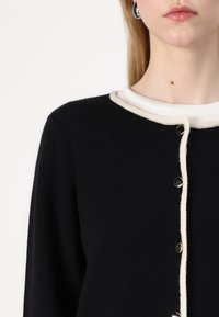 Black knitted cardigan featuring a cream-colored ribbed trim and button closure. Silver buttons accent the front, highlighting the textured fabric.