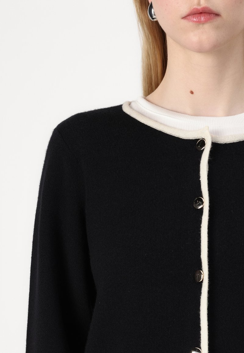 Cardigan nero lavorato a maglia con bordo in coste di colore crema e chiusura con bottoni. Bottoni argentati adornano la parte anteriore, mettendo in risalto il tessuto testurizzato.