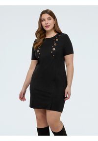 Mujer de pie con un vestido negro de manga corta con adornos circulares dorados, combinado con botas negras hasta la rodilla, sobre un fondo liso.