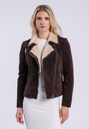 Modella femminile che indossa una giacca in suede marrone con un colletto fluffy, posando di fronte a uno sfondo neutro, mostrando un outfit alla moda.
