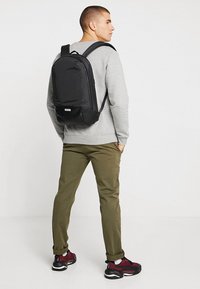 Zaino nero dal design elegante, realizzato in materiale resistente, con tasca frontale con zip e spallacci regolabili, posizionato su una felpa grigia.