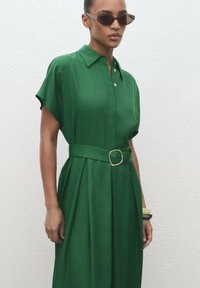 Green shirt dress with a collared neckline, short sleeves, a belted waist featuring a gold buckle, and a smooth, flowing fabric texture.