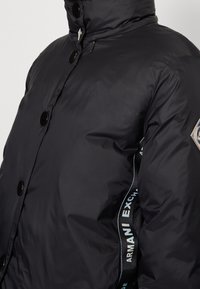 Mustat puffatakki, jossa on korkea kaulus, kiiltävät mustat napit ja sivumerkintä, jossa lukee "ARMANI EXCHANGE" valkoisella ja sinisellä tekstillä.