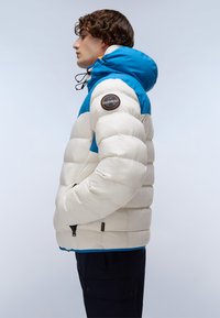 Ung man i vit och blå vadderad jacka med huva, händerna i fickorna, visad i vänsterprofil mot en enkel bakgrund.