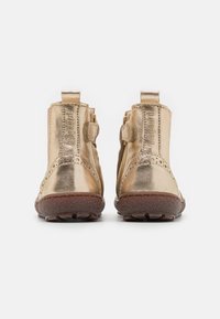 Bottes métalliques dorées à chevilles avec des détails de brogue, en cuir texturé, et une languette pour un enfilage facile. Semelles en caoutchouc avec des caractéristiques de traction pour une bonne adhérence.