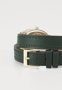 Bracelet de montre en cuir vert avec quincaillerie dorée, présentant un cadran rond et des coutures détaillées. Ajustable avec plusieurs trous pour un meilleur ajustement.