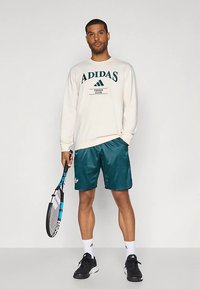 Sweatshirt creme claro com impressão verde "ADIDAS TENNIS CLUB", combinada com calções brilhantes verde-azulado e sapatos desportivos pretos, segurando uma raquete de ténis.