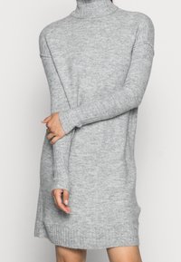 Robe-pull grise en maille avec un col roulé haut, des manches longues et des accents côtelés à l'ourlet, mettant en valeur un tissu doux et texturé.