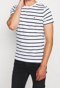 Mies, joka käyttää valkoista lyhythihaista T-paitaa, jossa on mustia vaakaraitoja ja pieni logo, yhdistettynä mustiin housuihin, käsi lähellä kasvojaan.