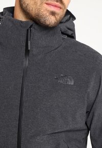 Gros plan d'un homme portant une veste à capuche gris foncé avec une fermeture éclair à l'avant et le logo "The North Face" sur la poitrine.