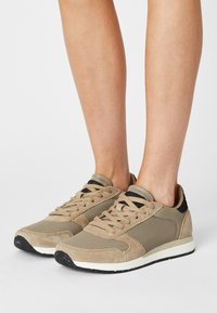 Beige tennarit, joissa on yhdistelmä verkko- ja suedettimateriaaleja, kumipohjat ja kangasnauhat, joissa on mustat yksityiskohdat kantapäässä.