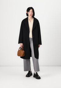 Μαύρο παλτό από μάλλινο ύφασμα με μεγάλα πέτα, κλείσιμο με κουμπί, μπεζ πουλόβερ από κάτω, γκρι τζιν καμπάνα και μια μικρή καφέ τσάντα με υφή.