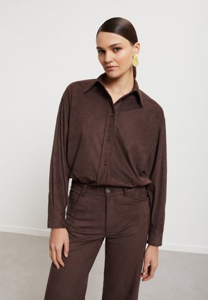 Vrouw met naar achteren gestylede haren draagt een bruin blouse met knopen en bijpassende broek, geaccentueerd met grote gouden ronde oorbellen.