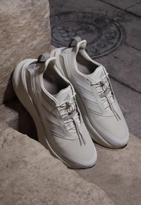 Coppia di sneakers bianche Adidas con tre strisce, lacci e suole spesse posate su gradini di pietra vicino a un tombino di metallo.