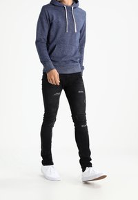 Felpa con cappuccio blu navy con coulisse grigio chiaro, abbinata a jeans skinny neri strappati e sneakers bianche con suola in gomma.