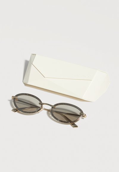Οβάλ σκουρόχρωμα γυαλιά ηλίου με λεπτά χρυσά σκελετά και διακοσμητικές στρας δίπλα σε λευκή γωνιώδη σκληρή θήκη για γυαλιά ηλίου.