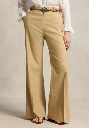 Beige bukser med vide ben og forlommer, kombineret med en hvid langærmet skjorte, brunt læderbælte og åbne sandaler.