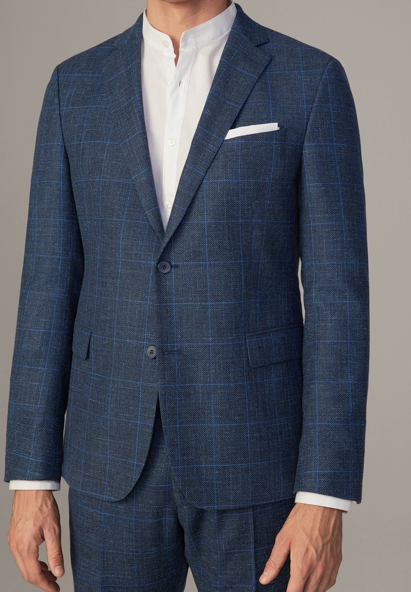 Homme portant un costume à carreaux bleu avec une chemise blanche à col droit et un mouchoir blanc dans la poche.