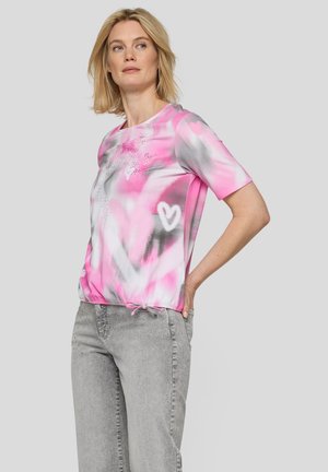 Frau trägt ein kurzärmeliges pink-weißes Batikshirt mit Herzmustern und hellgraue Jeans, steht mit einer Hand in der Hüfte.