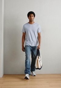 Tricou gri cu dungi, blugi albaștri și adidași albi; ținând o geantă de tote bej cu mânere închise; mediu interior cu podea din lemn.