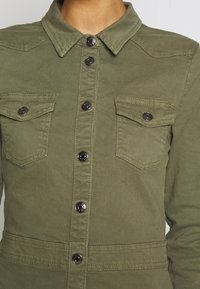 Chaqueta verde oliva con cierre de botones en la parte delantera, dos bolsillos en el pecho y cuello tipo solapa. Presenta botones de metal negro y una tela texturizada.
