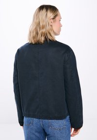 Marinblå jacka med en strukturerad design, med en synlig söm längs ryggen och långa ärmar. Tillverkad av ett slätt, enfärgat tyg.