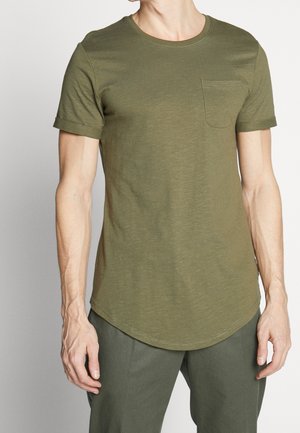 Homme portant un t-shirt à manches courtes olive avec col rond et poche poitrine, associé à un pantalon olive assorti, debout devant un fond uni.