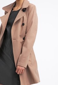 Kvinde iført en beige dobbeltradet frakke over en sort kjole med lynlås, stående foran en enkel hvid baggrund.