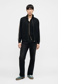 Nuori mies, jolla on musta vetoketjutakki, jossa on beigejä raitoja hihoissa, musta paita ja housut, seisomassa yksinkertaisen valkoisen taustan edessä.