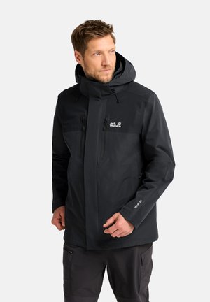 Man draagt een zwarte Jack Wolfskin waterdichte jas met capuchon, staat en kijkt opzij tegen een effen achtergrond.