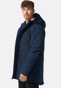 Mørkeblå parka med hætte, der har sorte detaljer, flere lommer og elastiske snore. Fremstillet af et slidstærkt materiale med en glat tekstur.