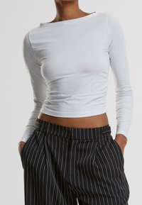 Weißes, langärmliges, kurzes Oberteil mit einem Boat-Neck-Ausschnitt, aus glattem Stoff, kombiniert mit schwarz gestreiften, hoch taillierten Hosen.