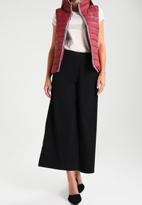 Femme portant un gilet matelassé rouge sans manches sur un t-shirt beige imprimé, un pantalon noir àLarge jambes et des escarpins noirs à bout pointu sur fond blanc.
