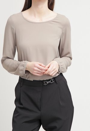 Kobieta w taupe bluzce z długim rękawem z marszczonymi mankietami i czarnych spodniach z wysokim stanem z paskiem z srebrną klamrą.