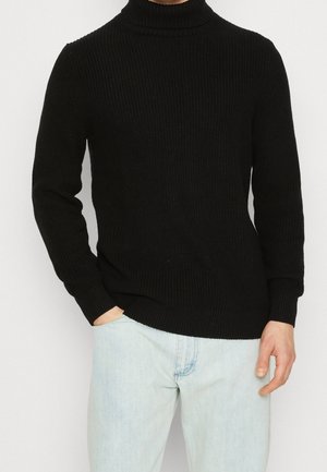 Schwarzer gerippter Rollkragenpullover mit langen Ärmeln, aus einem strukturierten Strickstoff, kombiniert mit hellblauen Jeans, die Hände in den Taschen.