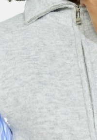 Pelēks, ar rāvējslēdzēju aizverams veste, izgatavota no mīkstas, teksturētas auduma. Iespējas stāvošs apkakle un sudraba rāvējslēdzējs. Redzama šūšanas detaļa gar šuvēm.