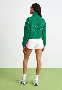 Mulher com cabelo trançado vestindo uma jaqueta verde com fecho, calções deportivos brancos e rosas, meias brancas e ténis prateados, em pé num chão verde.