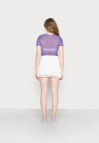 Top justo transparente roxo com uma textura ondulada, combinado com uma saia branca com babados. Modelo usa sapatos cinzas. Vista traseira exibida.