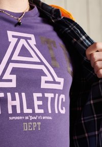 Fialové bavlněné tričko se velkým bílým a šedým grafickým textem "ATHLETIC" a "DEPT", pod kterým je položena kostkovaná košile.