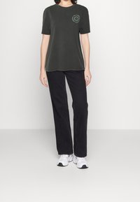 Camiseta de algodón verde oscuro con mangas cortas, escote redondo, que presenta un diseño gráfico circular en el lado izquierdo del pecho. Combinada con pantalones negros.