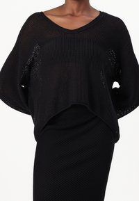 Pull noir en maille avec une coupe ample, un col en V et des manches larges. Présente un motif texturé et un ourlet raccourci, associé à une jupe noire.