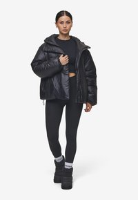 Zwarte pufferjack met een capuchon, glanzende textuur en drukknoopsluitingen. Draag het over een zwart crop top en leggings, gecombineerd met zwarte winterlaarzen.