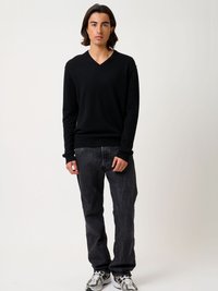 Schwarzer V-Ausschnitt-Pullover aus weichem Stoff, kombiniert mit dunklen Jeans. Das Model trägt weiße und graue Sneakers und steht vor einem schlichten Hintergrund.
